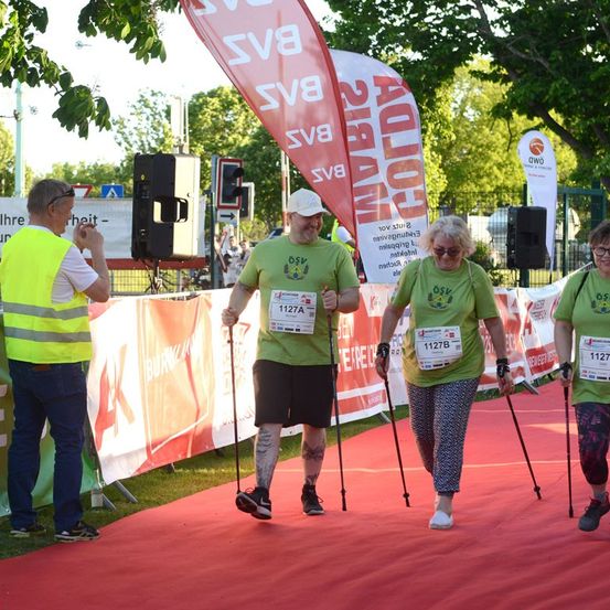 Drei Personen mit Gehstöcken laufen auf einem roten Teppich, wahrscheinlich an einem Marathon teilnehmend. Sie tragen grüne Shirts mit Nummern. Ein Mann in einer Warnweste steht in der Nähe.
