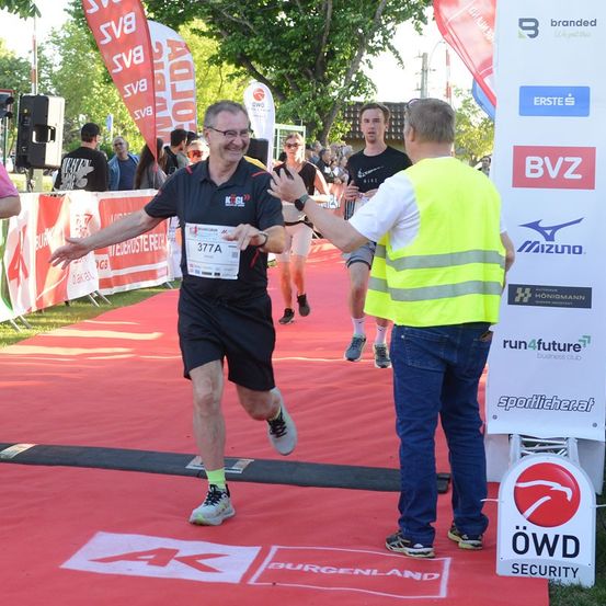 Ein Mann in einem schwarzen Shirt mit der Nummer 377A auf der Brust läuft auf einem roten Teppich bei einem Marathon. Ein Mann in einer gelben Weste schüttelt ihm die Hand.