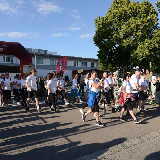Eine Gruppe von Menschen läuft an einem sonnigen Tag bei einem Marathon. Sie tragen weiße Hemden und schwarze Shorts. Einige von ihnen halten Stöcke. Hinter ihnen steht ein Gebäude mit einem roten Banner.