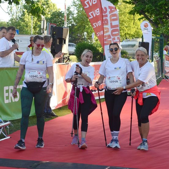Vier Frauen mit Laufausrüstung und Startnummern stehen auf einem roten Teppich, lächeln und posieren für ein Foto. Dahinter hält ein Mann ein Mikrofon und ein Banner mit Text.
