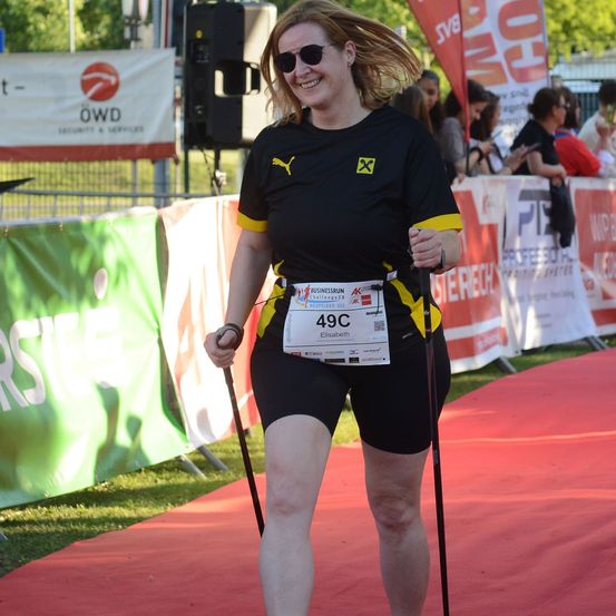 Eine Frau mit einer schwarzen und gelben Jacke, auf der die Nummer 49C steht, läuft einen Marathon. Sie hält Skistöcke und lächelt. Hinter ihr stehen Banner und Menschen.