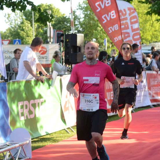 Zwei Läufer nehmen an einem Marathon teil, einer trägt ein rotes Shirt mit der Nummer 316C, der andere ein schwarzes Shirt mit der Nummer 32C. Sie laufen auf einem roten Teppich mit Bannern und Zuschauern im Hintergrund.