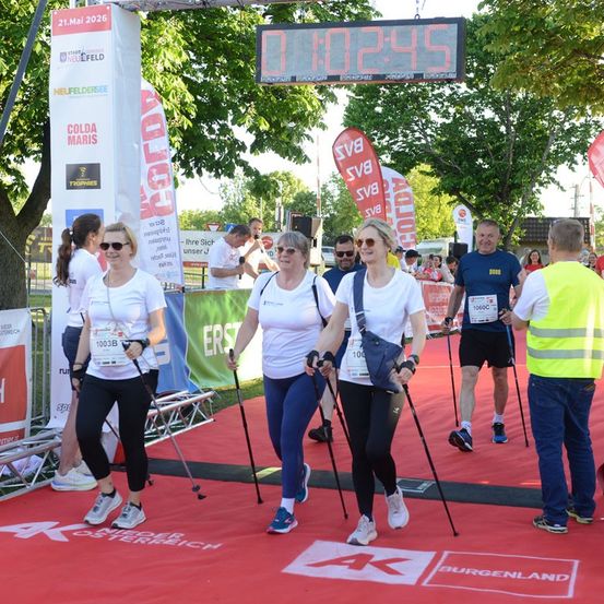 Eine Gruppe von Menschen nimmt an einem Marathon teil und benutzt Nordic-Walking-Stöcke. Sie laufen auf einem roten Teppich, über dem ein digitaler Timer angebracht ist.