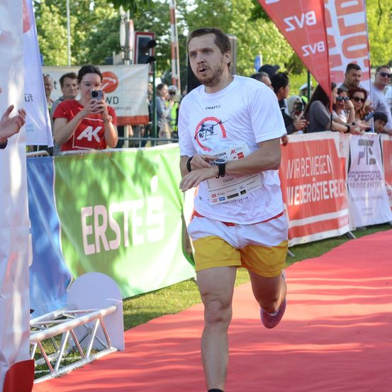 Ein Mann läuft in einem Marathon in einem weißen T-Shirt mit Logo und gelben Shorts. Zuschauer schauen von hinter einer Barriere zu, an der ein Banner mit Erstee steht.