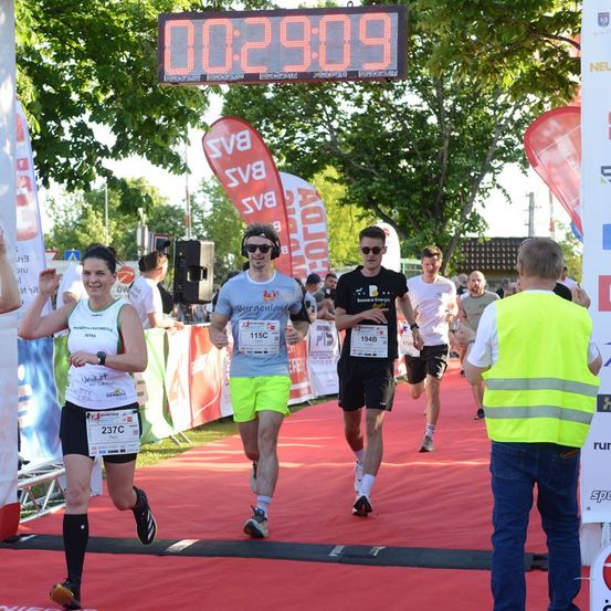 Läufer nehmen an einem Marathon teil, wobei eine digitale Uhr über ihnen die Zeit 00:29:09 anzeigt. Eine Frau in Weiß und Schwarz führt, gefolgt von einem Mann in Blau und Gelb. Zuschauer und Offizielle sind anwesend.