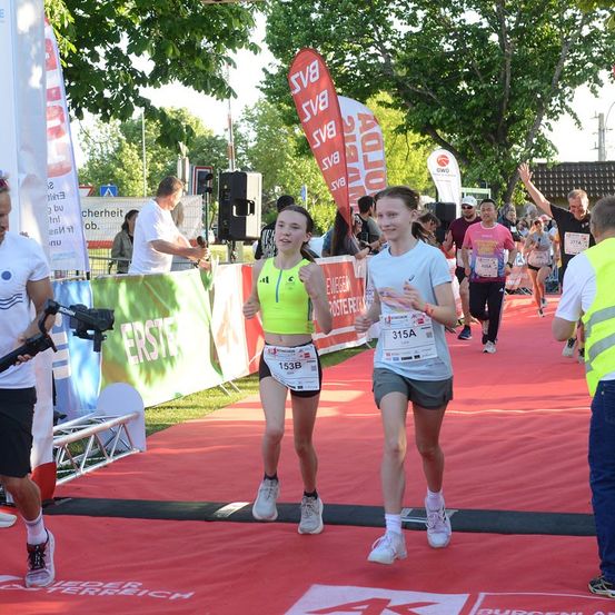 Zwei junge Frauen laufen auf einem roten Teppich bei einem Marathon-Event. Sie tragen Laufnummern und werden von einer Menge Läufer verfolgt. Im Hintergrund befinden sich Banner und ein Lautsprecher.