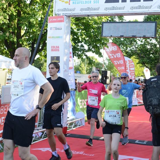 Eine Gruppe von Läufern, darunter ein Mann in einem weißen T-Shirt, ein Mann in einem schwarzen T-Shirt, ein Mann in einem rosa T-Shirt und ein junges Mädchen in einem grünen T-Shirt, laufen bei einem Marathon.