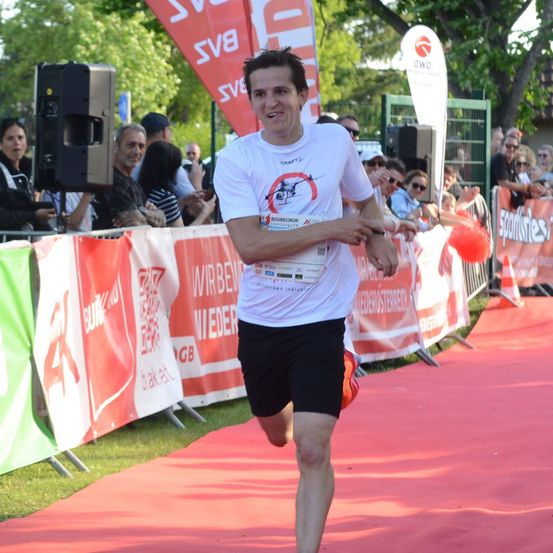 Ein Mann in einem weißen T-Shirt mit rotem Logo läuft auf einem roten Teppich bei einem Marathon, mit Zuschauern hinter einem Zaun, die zusehen.