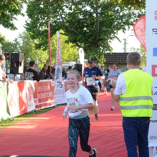 Ein junges Mädchen läuft auf einem roten Teppich während eines Marathons, während ein Mann in einer Warnweste sie beobachtet. Andere Läufer und Zuschauer sind im Hintergrund.