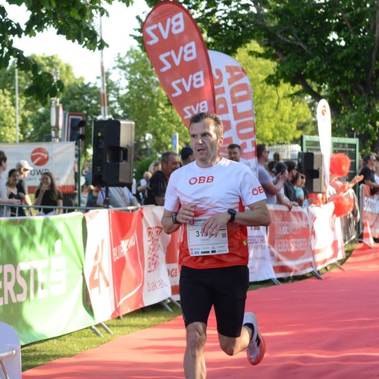 Ein Mann läuft bei einem Marathon in einem weißen Shirt mit der Nummer 3 und einer orangefarbenen Weste. Er ist auf einem roten Teppich mit Bannern und Zuschauern hinter ihm.