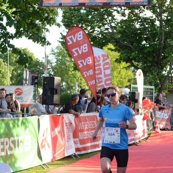 Ein Läufer in einem blauen Shirt und schwarzen Shorts läuft auf einem roten Teppich bei einem Marathon, mit Zuschauern hinter einem Zaun.
