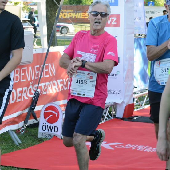 Ein älterer Mann mit einem pinken Shirt und einem Startnummernband läuft auf einem roten Teppich mit einem Banner im Hintergrund, das 'Bewegen der Storre' steht.