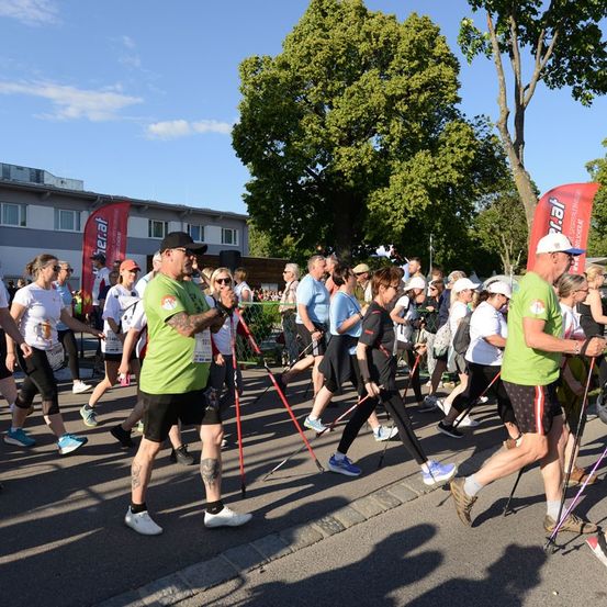 Eine Gruppe von Menschen läuft bei einem Marathon, einige benutzen Gehstöcke. Sie tragen grüne Shirts und schwarze Shorts. Im Hintergrund sind Bäume und Gebäude zu sehen.