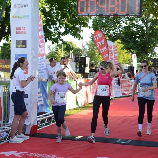 Eine Marathonläuferin, die ein rosa Shirt und eine Sonnenbrille trägt, wird von einer Frau und einem jungen Mädchen beglückwünscht. Die Marathonläuferin hat die Startnummer 10068. Sie laufen auf einem roten Teppich.