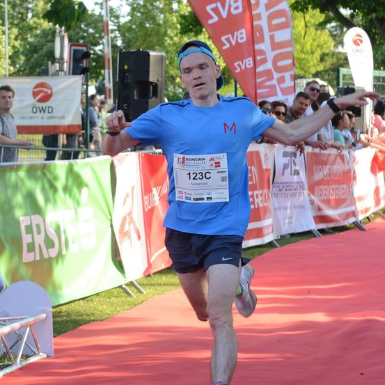 Ein männlicher Läufer in einem blauen Shirt und schwarzen Shorts läuft auf einem roten Teppich bei einem Marathon. Er hat einen Nummernchip auf seinem Shirt. Hinter ihm befinden sich Banner mit Text und Logos.