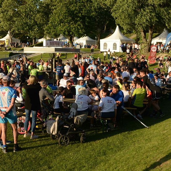 Eine große Menschenmenge versammelt sich in einem Park, einige sitzen an Picknicktischen, andere stehen, mit Zelten und einer Bier-Bar im Hintergrund.
