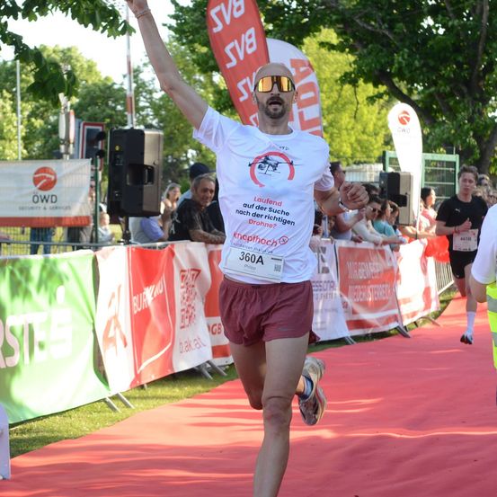 Ein Mann in einem weißen T-Shirt mit rotem Logo und roten Shorts läuft auf einem roten Teppich während eines Marathons, mit einer Menge, die hinter einem Zaun zusieht.