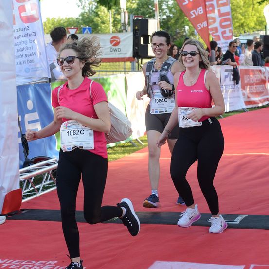 Drei Frauen in pinker und schwarzer Sportbekleidung laufen auf einem roten Teppich bei einem Marathon-Event. Die Frau vorne hat ein Nummernschild an ihrem Shirt. Dahinter befinden sich Banner und eine Menge Menschen.