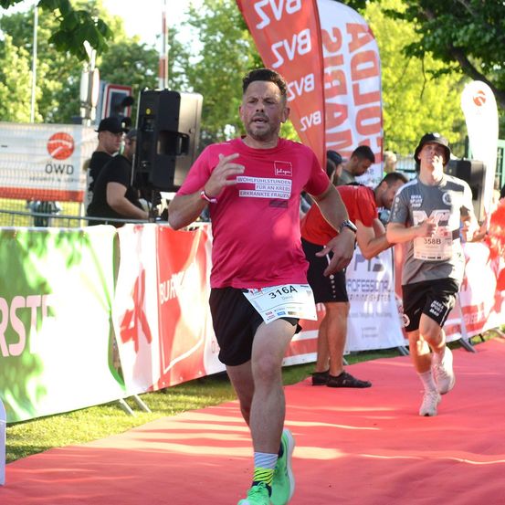 Ein Mann in einem roten T-Shirt und schwarzen Shorts läuft auf einem roten Teppich während eines Marathons. Hinter ihm ist ein weiterer Läufer zu sehen, und mehrere Personen stehen in der Nähe von Bannern und einem Lautsprecher.