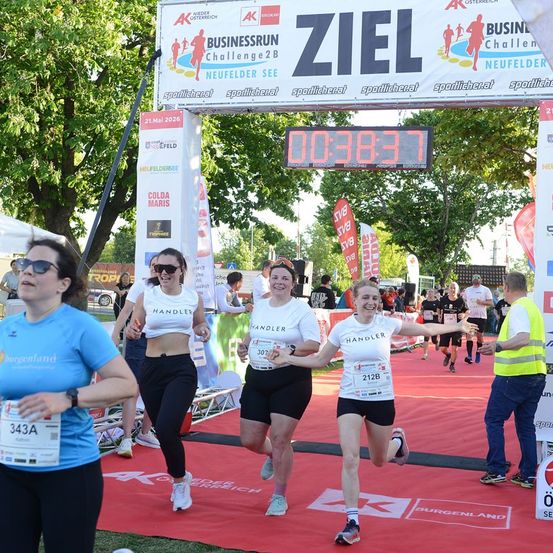 Drei Frauen in weißen Shirts und schwarzen Shorts laufen in einem Marathon. Sie halten Händchen und lächeln. Die Ziellinie ist im Hintergrund mit einem digitalen Timer, der 00:38:37 anzeigt.