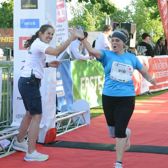 Zwei Frauen, eine mit einem blauen Shirt mit der Nummer 91B, geben sich gegenseitig einen High-Five bei einem Marathon-Event. Sie stehen auf einem roten Teppich mit einem Banner mit der Aufschrift 'Erste' im Hintergrund.