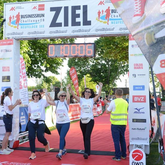 Drei Frauen in weißen Shirts und blauen Hosen feiern das Überqueren der Ziellinie eines Marathons. Ein Mann in einer gelben Weste steht in der Nähe.