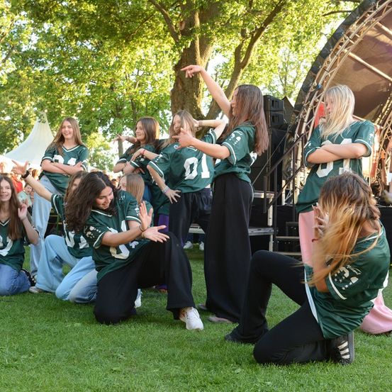 Eine Gruppe junger Frauen in grünen Trikots führt eine Tanzroutine auf einem Rasenfeld auf, mit einer Bühne und Lautsprechern im Hintergrund.