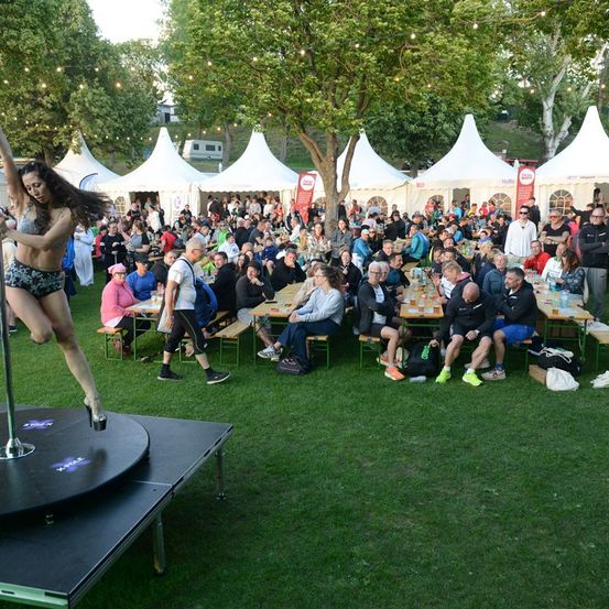 Eine Frau führt eine Pole-Tanz-Darbietung vor einer Menschenmenge bei einer Outdoor-Veranstaltung mit weißen Zelten und Tischen auf.