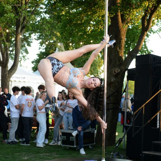Eine Frau führt eine Poledance-Darbietung im Freien durch, trägt einen blauen Bikini und hohe Schuhe, während eine Menschenmenge zuschaut.