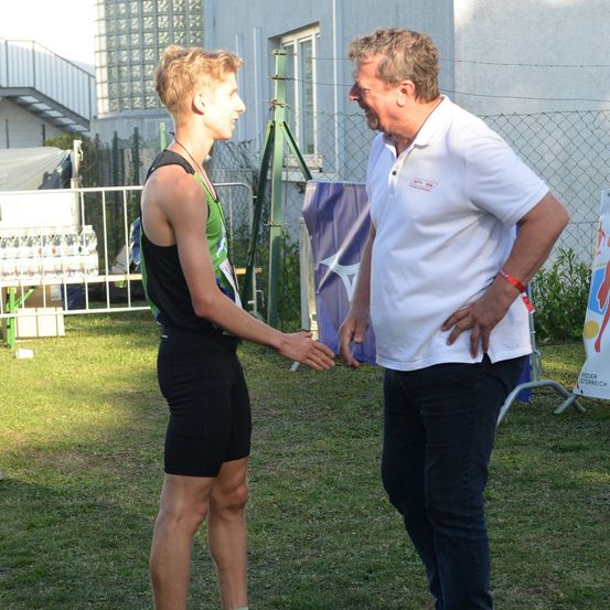 Ein Mann in einem grünen und schwarzen Sportoutfit schüttelt die Hand eines älteren Mannes in einem weißen Poloshirt. Sie stehen auf einem Rasenfeld mit einem Zaun und einem Banner im Hintergrund.