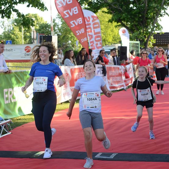 Drei Läufer, darunter eine Frau und zwei Mädchen, laufen bei einem Marathon mit einem roten Teppich und Bannern im Hintergrund.