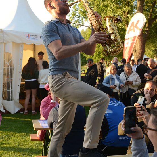Ein Mann spielt Saxophon auf einer Bühne im Park, während eine Menge Menschen zuschaut und einige Fotos machen.