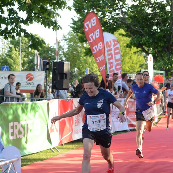 Eine Läuferin mit der Nummer 272B auf dem Trikot läuft bei einem Marathon. Zuschauer beobachten sie hinter einer Absperrung. Ein weiterer Läufer folgt ihr.