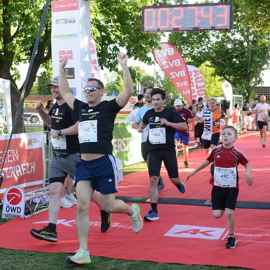 Teilnehmer eines Marathons laufen auf einem roten Teppich, über ihnen zeigt eine digitale Uhr die Zeit an. Bäume und Banner säumen die Seiten.