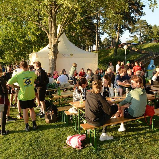 Eine Gruppe von Menschen versammelt sich bei einer Outdoor-Veranstaltung, einige sitzen an Tischen, während andere herumstehen, mit einem großen weißen Zelt im Hintergrund.