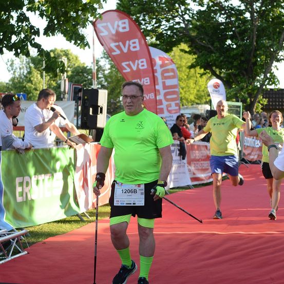 Ein Mann in einem neongrünen Shirt und schwarzen Shorts läuft einen Marathon und hält einen Gehstock. Er hat die Nummer 1206B auf seinem Shirt. Hinter ihm nehmen auch zwei andere Läufer am Marathon teil.