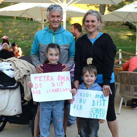 Eine vierköpfige Familie steht lächelnd zusammen, wobei zwei Kinder Schilder halten. Das Mädchen hält ein Schild mit der Aufschrift 'Petra Oma, du bist die schnellste' und der Junge hält ein Schild mit der Aufschrift 'Rudi op, du bist der schnellste'. Hinter ihnen sitzen und stehen Leute unter Schirmen.