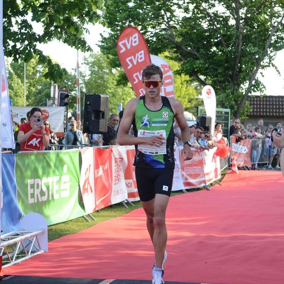 Ein männlicher Athlet in einem grünen und schwarzen Sportoutfit läuft auf einem roten Teppich bei einem Marathon-Event, mit Zuschauern und Bannern im Hintergrund.