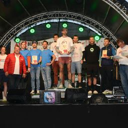 Eine Gruppe von Männern steht mit Trophäen auf der Bühne, ein Mann in der Mitte trägt einen Hut und hält eine große Trophäe.