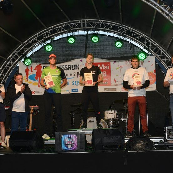 Fünf Männer stehen auf einer Bühne und halten Urkunden in der Hand, zwei mit grünen Shirts, einer mit einer roten Kappe und zwei in schwarzen Shirts. Dahinter hängt ein Banner mit einer Lauffigur und Text. Musikinstrumente und Lautsprecher stehen auf der Bühne.