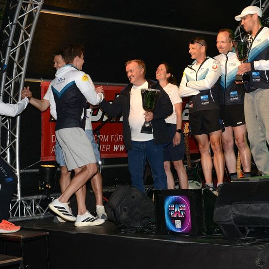 Eine Gruppe von Menschen steht auf einer Bühne, hält Trophäen und lächelt. Sie tragen Sportbekleidung und Turnschuhe. Ein Banner mit rotem Text steht hinter ihnen, und ein Lautsprecher befindet sich auf der Bühne.