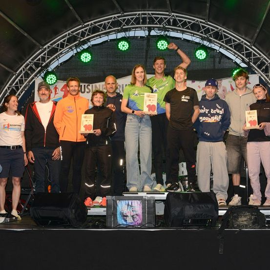 Eine Gruppe von Menschen steht auf einer Bühne und hält Auszeichnungen in der Hand, mit grünen Lichtern und einem Banner im Hintergrund.