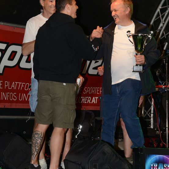 Ein Mann in einer schwarzen Kapuzenjacke und grünen Shorts schüttelt die Hand eines Mannes in einem weißen T-Shirt und blauen Jeans, der einen Pokal hält. Dahinter ist ein Banner mit rotem Text und ein dritter Mann in einem weißen T-Shirt und Jeansshorts.