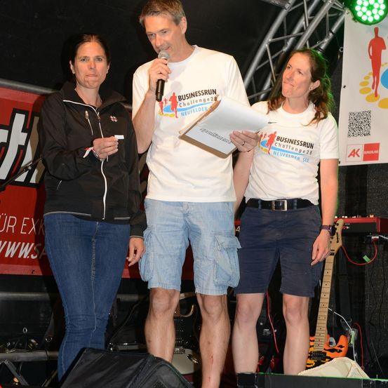 Drei Personen stehen auf einer Bühne, ein Mann hält ein Mikrofon und liest von einem Blatt Papier, während zwei Frauen neben ihm stehen, eine hält ein Blatt Papier. Hinter ihnen ist ein Banner mit Text und eine Gitarre.
