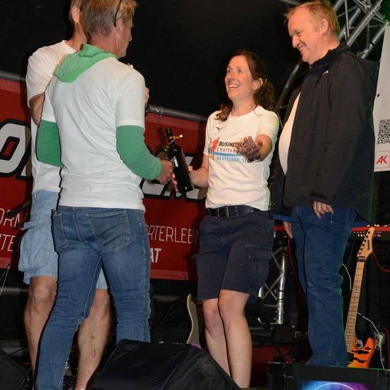 Eine Frau in einem weißen Shirt mit Logo steht auf der Bühne mit drei Männern, von denen einer eine Flasche hält. Sie alle lächeln. Dahinter ist ein roter Hintergrund mit Text und einer Gitarre.