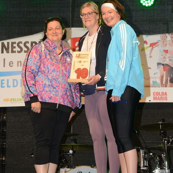 Drei Frauen stehen auf einer Bühne, eine hält eine Plakette mit der Nummer 10. Die Frau auf der linken Seite trägt eine bunte Jacke, die mittlere Frau trägt eine Brille und eine Stirnband, und die rechte Frau trägt eine blaue Jacke. Hinter ihnen ist ein Banner mit dem Text 'NESS' und 'ELD'.