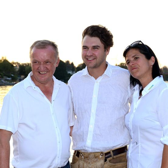Drei Menschen stehen zusammen in der Nähe eines Gewässers und lächeln für ein Foto. Der Mann links ist älter, der Mann in der Mitte ist jung und die Frau rechts hat eine Sonnenbrille auf dem Kopf.