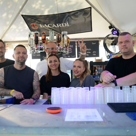 Eine Gruppe von Menschen posiert für ein Foto an einer Bar mit Bacardi-Marke. Sie lächeln und stehen hinter einer Theke mit Gläsern und einem Ventilator.
