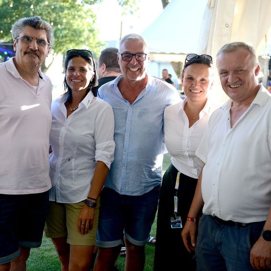 Fünf Menschen stehen zusammen auf einem Rasen und lächeln für ein Foto. Sie sind lässig gekleidet und befinden sich wahrscheinlich bei einer gesellschaftlichen Veranstaltung.