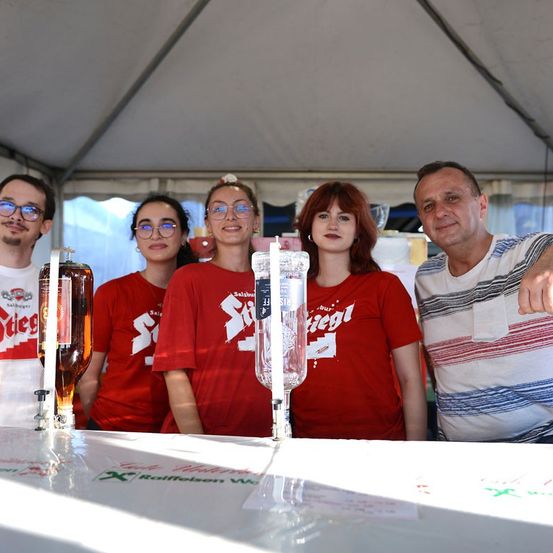 Fünf Personen in roten T-Shirts stehen hinter einem Tisch mit Bierflaschen und Gläsern, möglicherweise auf einem Festival.